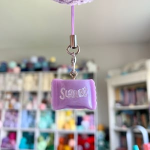 May include: A lavender-colored candy charm with the word "Sweet" in white script. The charm is attached to a silver clasp and a thin lavender cord, hanging from a larger, fuzzy, lavender object.