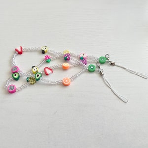 May include: A white beaded phone charm with colorful fruit charms, including strawberries, pineapples, kiwis, and oranges. The charm has two white cords attached.