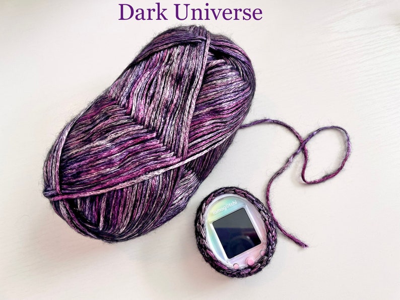 Pode incluir: Uma bola de fio roxo e preto com um brilho prateado, etiquetada como "Dark Universe". Uma pequena bolsa cinza de croch&ecirc; com um brinquedo digital para animais de estima&ccedil;&atilde;o dentro.
