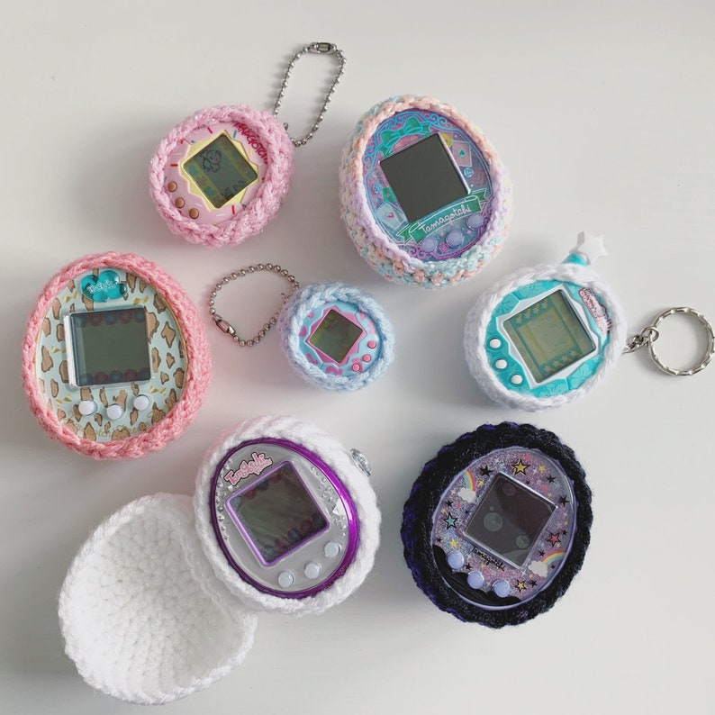 Pode incluir: Uma cole&ccedil;&atilde;o de nove chaveiros Tamagotchi de croch&ecirc; coloridos. Cada chaveiro apresenta um design diferente, incluindo um chaveiro rosa e branco com um design de cora&ccedil;&atilde;o, um chaveiro azul e branco com um design de estrela e um chaveiro roxo e branco com um design de flor.