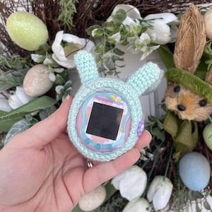 Puede incluir: Una funda de ganchillo de color azul claro en forma de conejo para una mascota digital Tamagotchi rosa y azul. La funda tiene una flor morada y botones azules. El Tamagotchi está en la funda y la pantalla es negra.