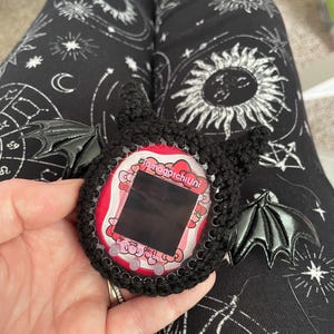 May include: Black crocheted  Tamagotchi Uni case with bat wings. The case has a pink and white screen with the text "Tamagotchi Uni" on it.