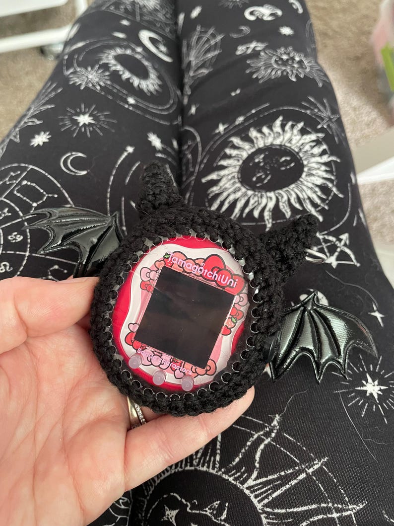 May include: Black crocheted  Tamagotchi Uni case with bat wings. The case has a pink and white screen with the text "Tamagotchi Uni" on it.