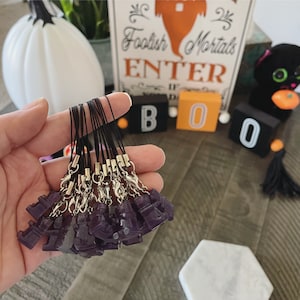 May include: A hand holds several small, dark purple castle charms attached to black cords with silver clasps. In the background, a white pumpkin, a sign that says "Foolish Mortals ENTER", and "BOO" block letters are visible.