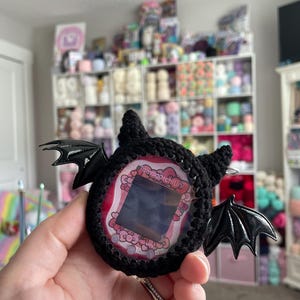 May include: A black crocheted Tamagotchi with bat wings and a pink and white screen. The text "Tamagotchi Uni" is on the screen.