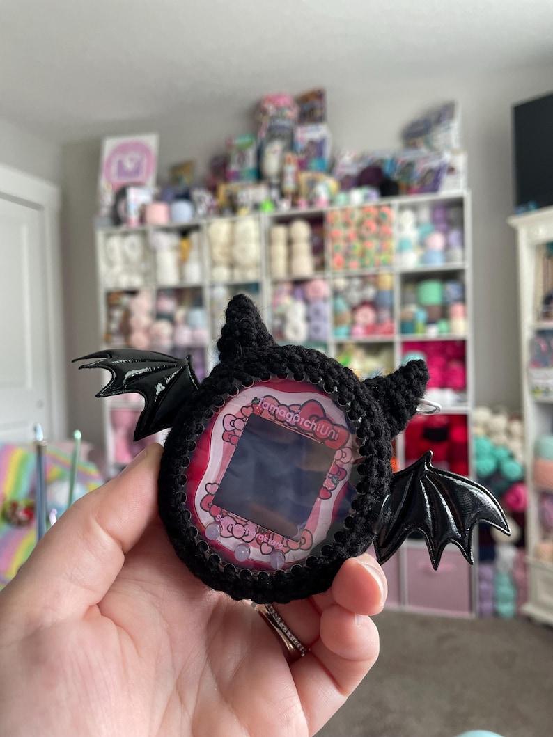 May include: A black crocheted Tamagotchi with bat wings and a pink and white screen. The text "Tamagotchi Uni" is on the screen.