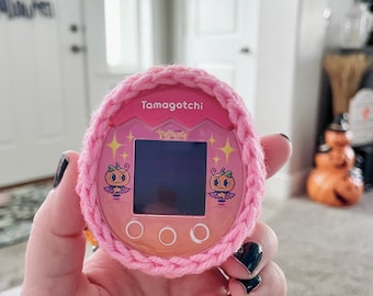 May include: A pink Tamagotchi with a crocheted cover. The screen displays a pumpkin character with the text "Happy Hallowen".