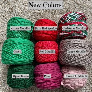Pode incluir: Nove novelos de fio em v&aacute;rias cores, incluindo verde, vermelho, ameixa e cinza. O fio est&aacute; disposto em tr&ecirc;s filas de tr&ecirc;s novelos cada. O fio est&aacute; etiquetado com os nomes das cores: Verde met&aacute;lico, Vermelho escuro brilhante, Met&aacute;lico natalino, Verde, Vermelho met&aacute;lico, Cinza met&aacute;lico, Verde alpino, Ameixa e Ouro rosa met&aacute;lico.