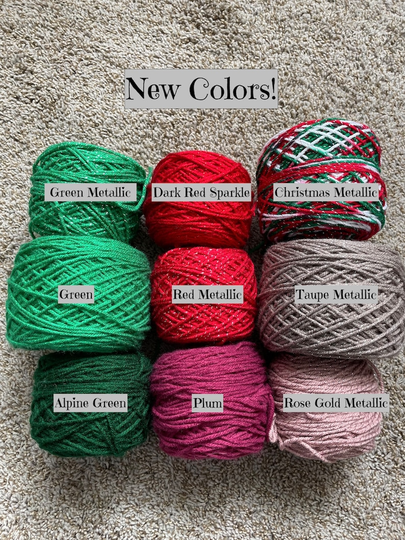 Pode incluir: Nove novelos de fio em v&aacute;rias cores, incluindo verde, vermelho, ameixa e cinza. O fio est&aacute; disposto em tr&ecirc;s filas de tr&ecirc;s novelos cada. O fio est&aacute; etiquetado com os nomes das cores: Verde met&aacute;lico, Vermelho escuro brilhante, Met&aacute;lico natalino, Verde, Vermelho met&aacute;lico, Cinza met&aacute;lico, Verde alpino, Ameixa e Ouro rosa met&aacute;lico.