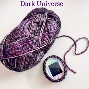 May include: A ball of purple, black, and white yarn with a silver sheen. The yarn is labeled "Dark Universe". A small, black, and silver electronic device with a screen is in the lower right corner of the image.