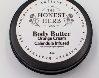 May include: A black tin container with a white label that reads "The Honest Herb Co." and "Body Butter Orange Cream Calendula Infused". The label also says "Made from simple organic ingredients" and "4 oz.". The tin is labeled with the words "smooths skin", "softens", "hydrates", and "for happy healthier skin".
