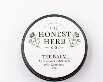 Könnte beinhalten: Ein schwarzes und weißes Etikett auf einem runden Blechbehälter lautet "The Honest Herb Co." und "The Balm All Purpose Herbal Salve With Calendula 2oz".