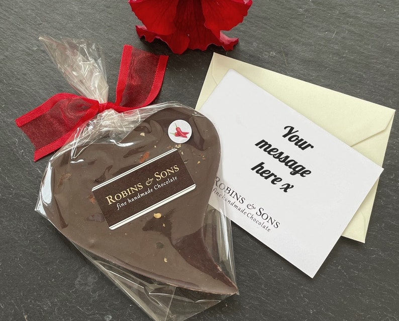 May include: A heart-shaped chocolate treat wrapped in clear cellophane and tied with a red ribbon. The chocolate has a label that reads "ROBINS & SONS fine handmade Chocolate". A card with the text "Your message here x" is included.