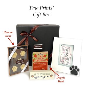 May include: A black gift box tied with a brown ribbon, containing dog-themed treats and a photo frame. Includes a box of paw-shaped chocolates, dog treats, and a wooden sign that reads "Be the person your dog thinks you are."