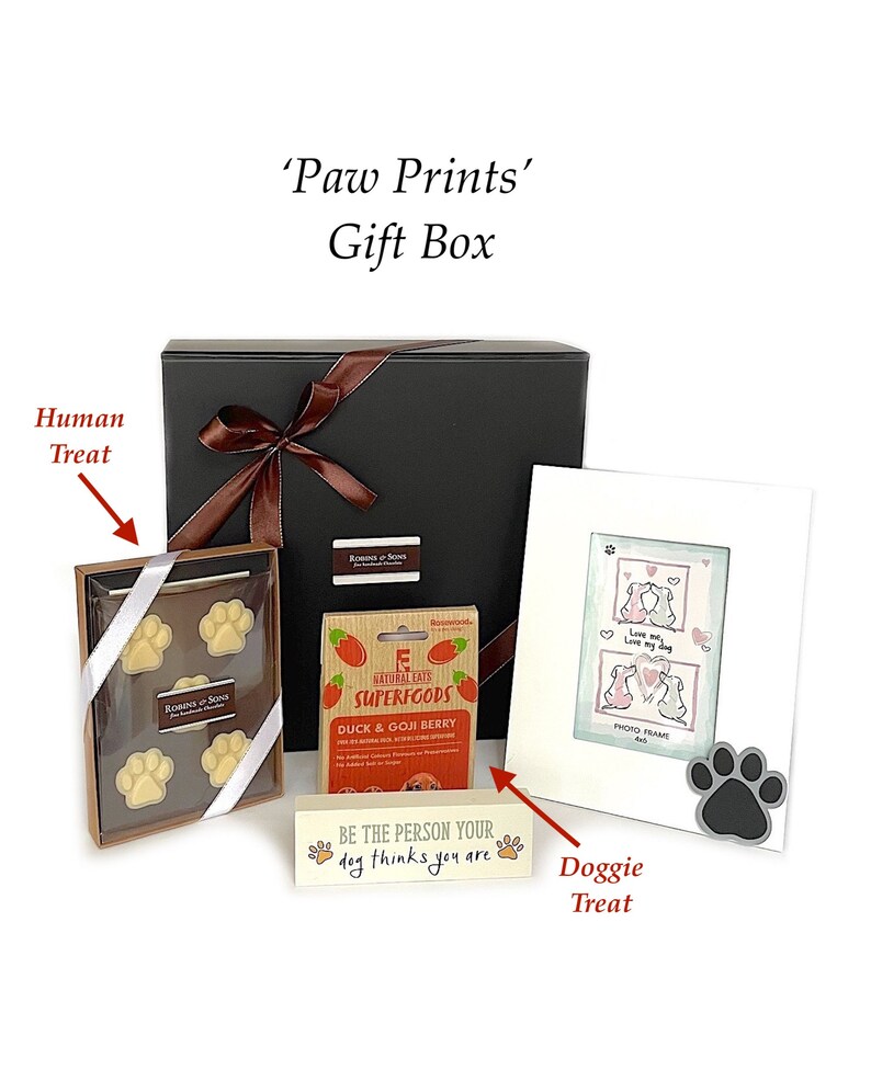 May include: A black gift box tied with a brown ribbon, containing dog-themed treats and a photo frame. Includes a box of paw-shaped chocolates, dog treats, and a wooden sign that reads "Be the person your dog thinks you are."