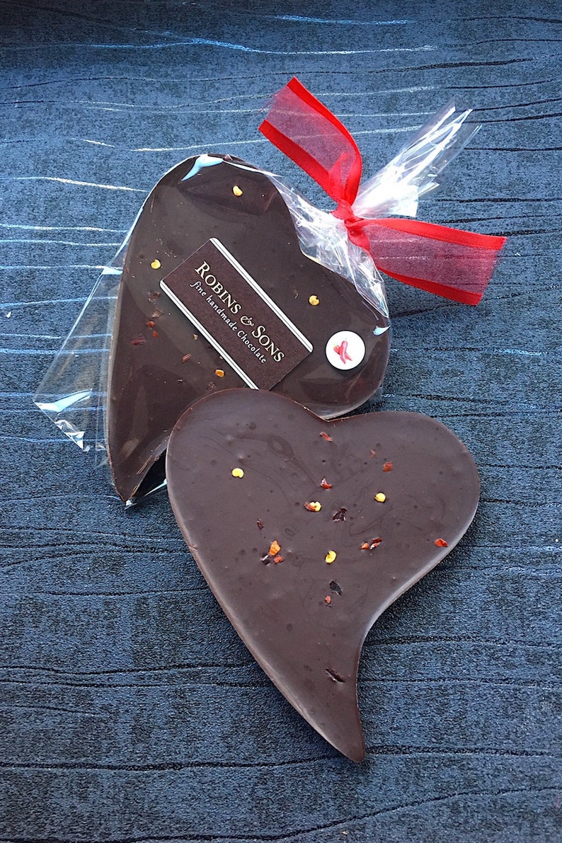 May include: Two heart-shaped dark chocolates, one unwrapped and one in clear cellophane tied with a red ribbon. The unwrapped chocolate has visible specks. The wrapped chocolate has a label that reads "Robins & Sons fine handmade Chocolate."