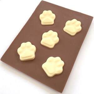 May include: A rectangular chocolate bar with five white chocolate paw prints arranged on top. The chocolate bar is a rich brown color, and the paw prints are a creamy white. The image is taken from a slightly elevated angle.
