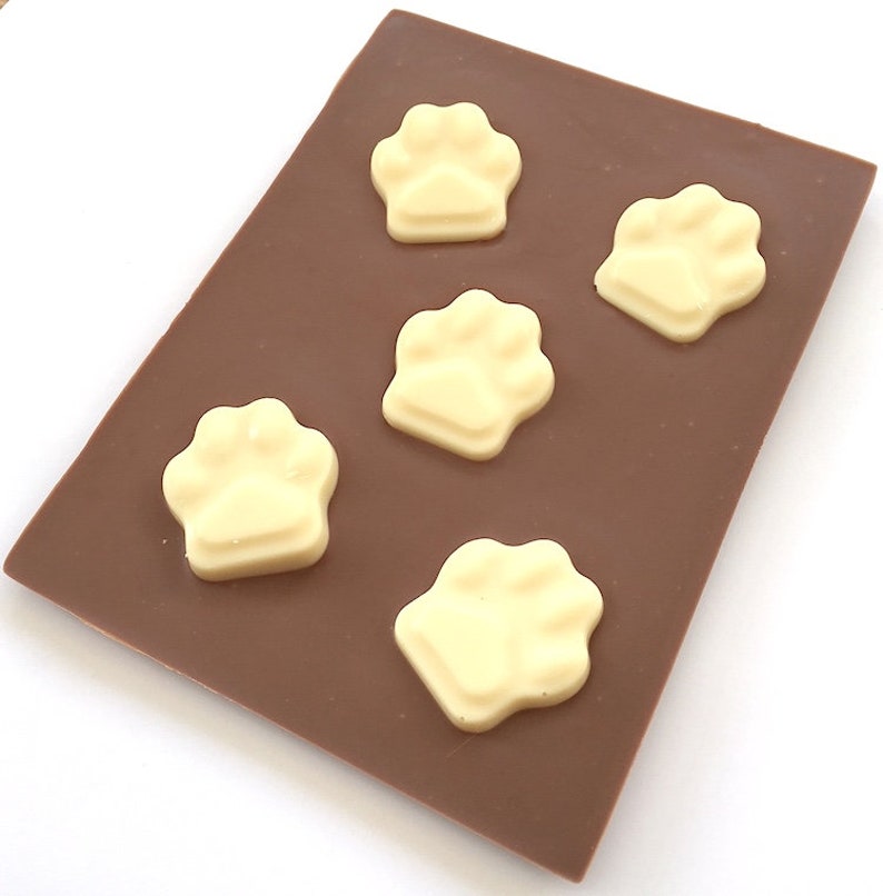 May include: A rectangular chocolate bar with five white chocolate paw prints arranged on top. The chocolate bar is a rich brown color, and the paw prints are a creamy white. The image is taken from a slightly elevated angle.