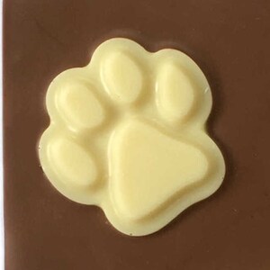 May include: A close-up of a light yellow paw print design on a dark brown surface. The paw print is raised and has a smooth texture. The background is a solid, dark brown color.