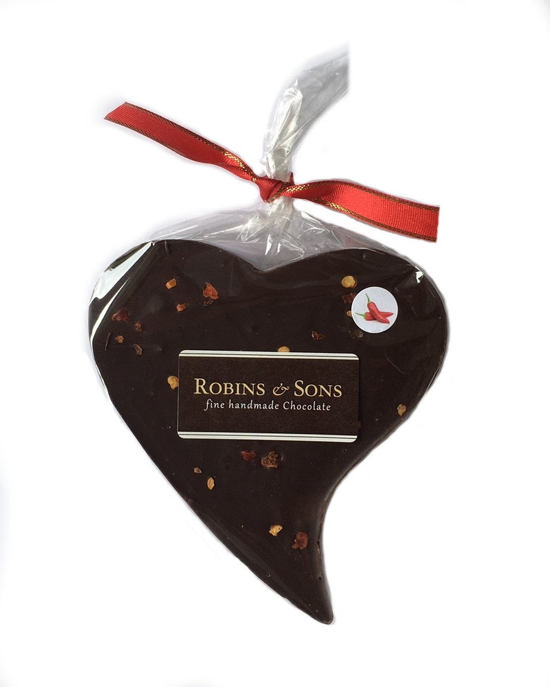 May include: A heart-shaped dark chocolate treat, sprinkled with visible flakes, is wrapped in clear cellophane and tied with a red ribbon. The chocolate has a sticker with a chili pepper image and a label that reads "Robins & Sons fine handmade chocolate."
