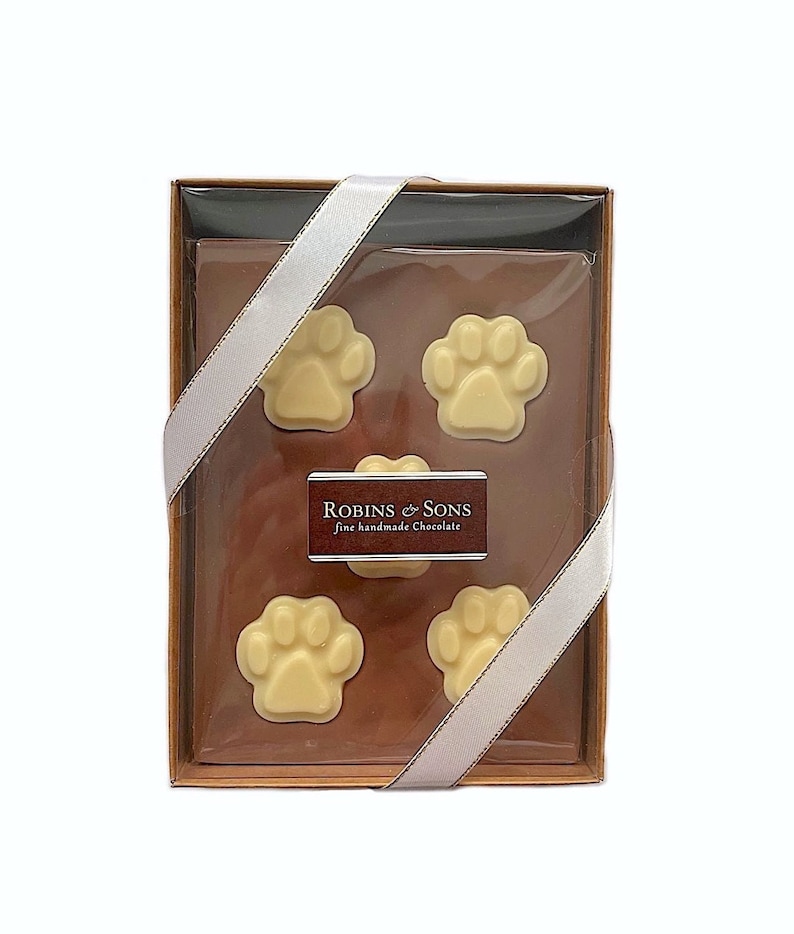 May include: A gift box of fine handmade chocolate from Robins & Sons. The box contains four paw-shaped chocolates in a light cream color, arranged on a brown background. The box is tied with a white ribbon.
