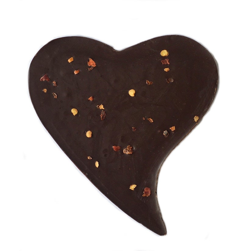 May include: A dark chocolate heart-shaped confection sprinkled with small, reddish-brown and yellow pieces. The chocolate has a smooth surface and a slightly irregular shape, set against a white background.