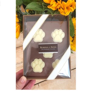 May include: A clear box contains four white chocolate paw prints, arranged on a brown surface. The box is tied with a white ribbon with gold trim. The label reads "Robins & Sons fine handmade Chocolate". Yellow sunflowers are in the background.