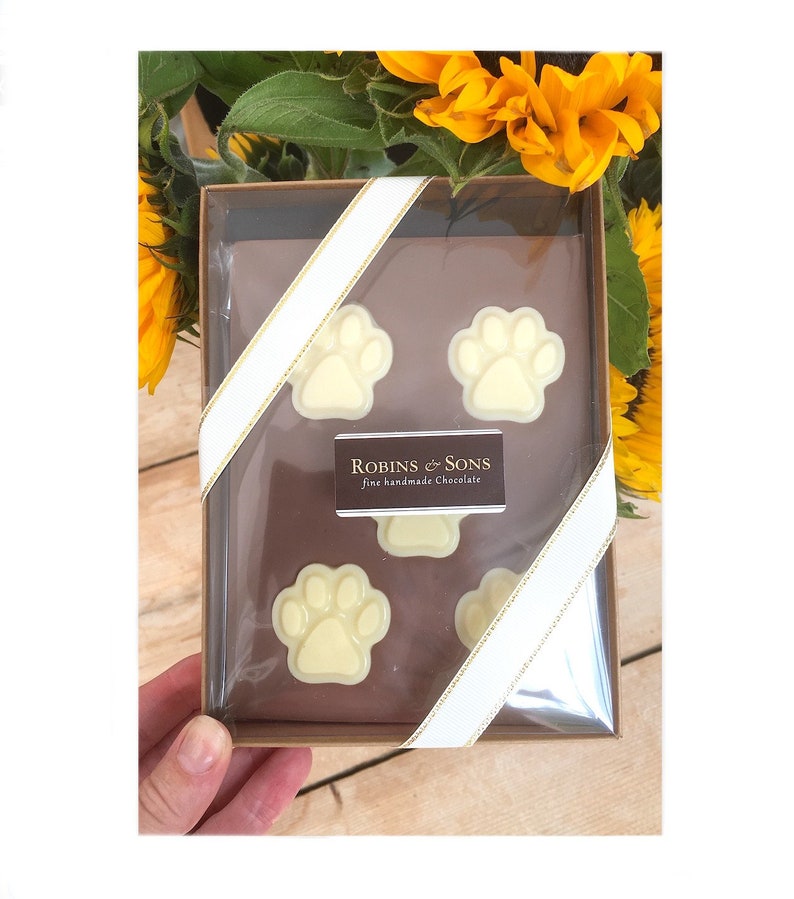 May include: A clear box contains four white chocolate paw prints, arranged on a brown surface. The box is tied with a white ribbon with gold trim. The label reads "Robins & Sons fine handmade Chocolate". Yellow sunflowers are in the background.