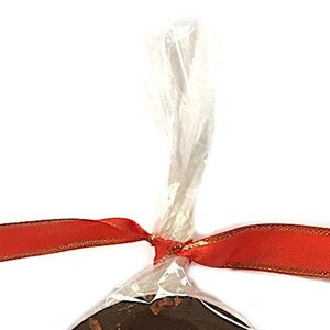 May include: A wrapped chocolate treat, tied with a red ribbon with gold trim. The clear cellophane wrapping is gathered at the top and tied with a bow. The background is plain white.