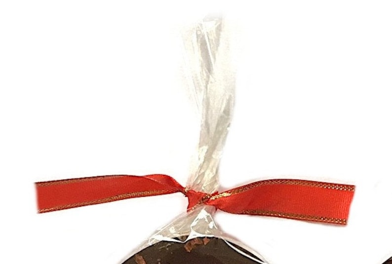 May include: A wrapped chocolate treat, tied with a red ribbon with gold trim. The clear cellophane wrapping is gathered at the top and tied with a bow. The background is plain white.