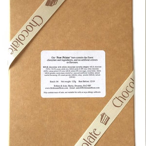 May include: A brown cardboard box tied with a cream ribbon printed with the word "Chocolate". The box contains a label with text about the ingredients of the "Paw Prints" chocolate bars. The ribbon has a small cake graphic.