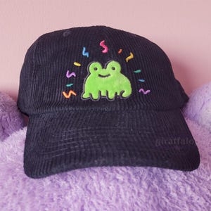 Può includere: Cappellino da baseball in velluto a coste nero con una rana verde ricamata sul davanti. La rana è circondata da coriandoli colorati.