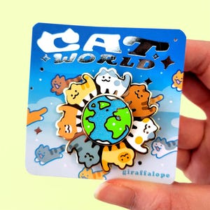 May include: A circular enamel pin featuring a central globe surrounded by eight stylized cats in various colors including orange, gray, white, and brown. The pin is attached to a blue backing card with the text CAT WORLD and giraffalope.