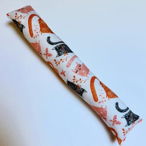 May include: A long, cylindrical fabric toy featuring a white background with a repeating pattern of orange and black cats, small pink butterflies, and delicate orange leafy sprigs. The cat figures are depicted in various poses.