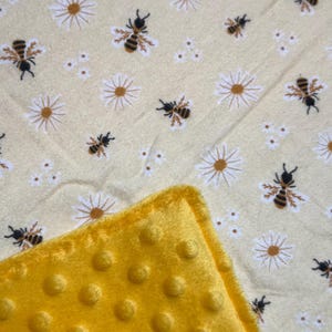 May include: A soft blanket featuring a light yellow fabric printed with a repeating pattern of small brown and black bees and white daisy flowers, paired with a textured bright yellow minky fabric with raised circular dimples on the corner.