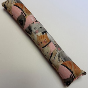 May include: A long, cylindrical fabric toy featuring a repeating pattern of illustrated cats in various colors including orange, gray, and black, set against a light background with pink accents and small heart motifs.