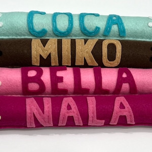 May include: Four felt dog bed rolls with paw prints and names embroidered on them. The names are Coca, Miko, Bella, and Nala. The rolls are in different colors: light blue, brown, pink, and purple.