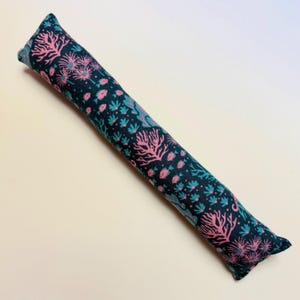 May include: A long, cylindrical fabric tube featuring a dark navy blue background with a repeating pattern of pink coral, teal sea anemones, and small pink fish. The textile object has a soft, stuffed appearance and is oriented diagonally.