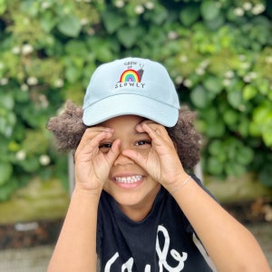 May include: A light blue baseball cap with a rainbow and snail graphic and the text "Grow Up Slowly". A child is wearing the cap and smiling.