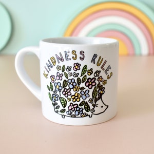 May include: White ceramic mug featuring a black and white illustration of a hedgehog with flowers on its back. The text "Kindness Rules" is written in a colourful font around the hedgehog.