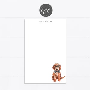 May include: A white notepad with the text "LUNA ROLDAN" printed at the top. A watercolor illustration of a brown dog wearing glasses and a bow tie is at the bottom of the notepad.
