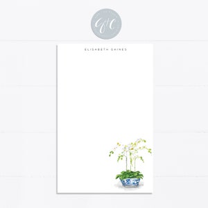 May include: White orchid flowers in a blue and white porcelain planter. The planter has a square shape with rounded corners. The flowers are arranged in a symmetrical pattern. The background is white.