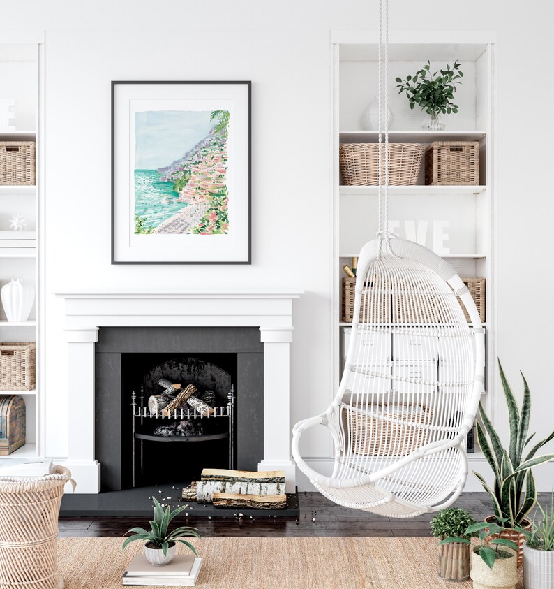 May include: A framed watercolor print of a coastal town hangs above a fireplace. A white wicker hanging chair is suspended from the ceiling. Wicker baskets and plants decorate the room, creating a bright and airy interior design.