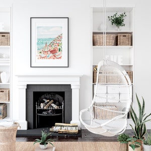 May include: A framed watercolor print of a coastal town with red-roofed buildings and a turquoise sea hangs above a fireplace. A white wicker hanging chair and built-in shelves with baskets and decor complete the bright, airy interior design.