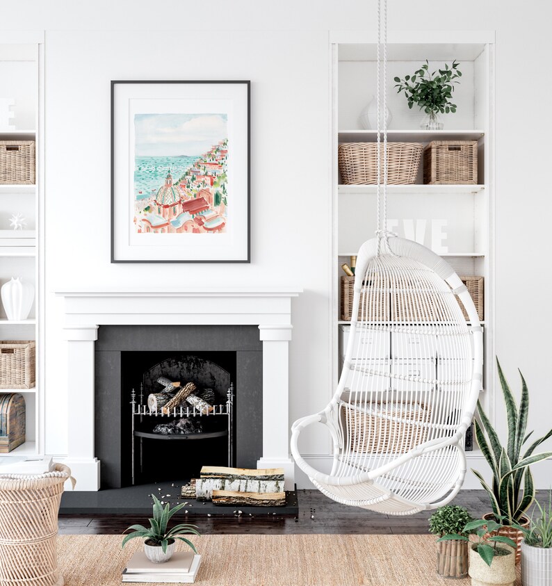 May include: A framed watercolor print of a coastal town with red-roofed buildings and a turquoise sea hangs above a fireplace. A white wicker hanging chair and built-in shelves with baskets and decor complete the bright, airy interior design.