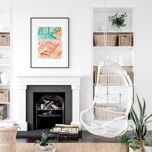 May include: A framed watercolor painting depicts a beach scene with red and orange beach chairs, turquoise water, and a green cliff. A white wicker hanging chair and built-in shelves with baskets and decor are also visible. The room has a fireplace and neutral color scheme.