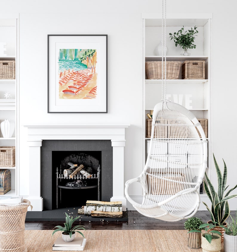 May include: A framed watercolor painting depicts a beach scene with red and orange beach chairs, turquoise water, and a green cliff. A white wicker hanging chair and built-in shelves with baskets and decor are also visible. The room has a fireplace and neutral color scheme.