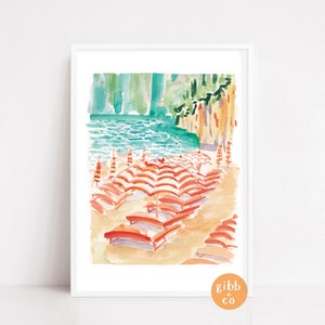 May include: Watercolor print of a beach scene with orange and white striped umbrellas and lounge chairs on a sandy shore. The artwork features turquoise water and a green cliffside in the background. The print is framed in a white border.