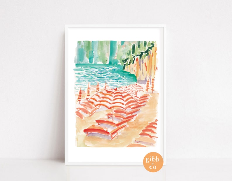 May include: Watercolor print of a beach scene with orange and white striped umbrellas and lounge chairs on a sandy shore. The artwork features turquoise water and a green cliffside in the background. The print is framed in a white border.