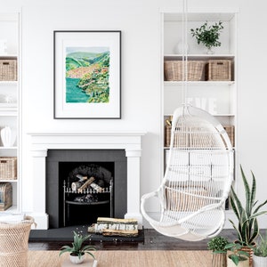 May include: A framed watercolor painting of a coastal town with green hills and turquoise water hangs above a fireplace. A white wicker hanging chair and built-in shelves with baskets and plants complete the cozy, bright interior.